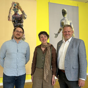 Eine Frau und zwei Männer vor zwei modernen Gemälden an einer gelben Wand.