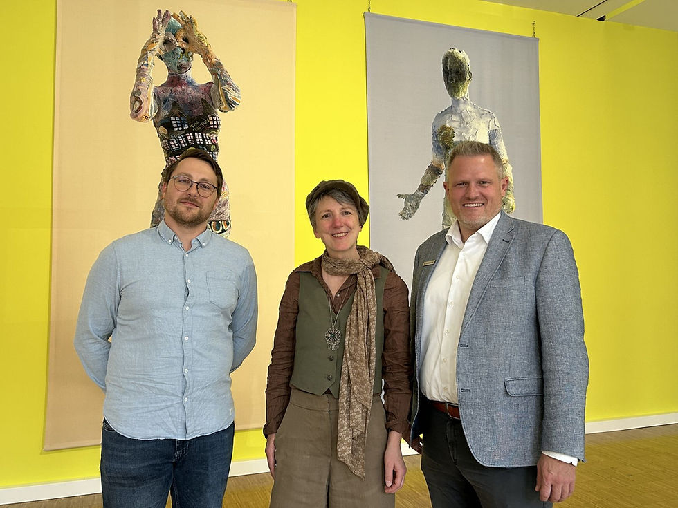 Eine Frau und zwei Männer vor zwei modernen Gemälden an einer gelben Wand.