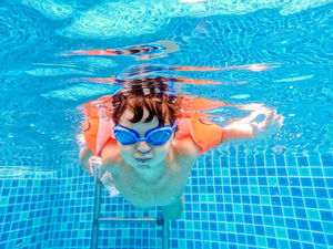 Junges Kind mit Schwimmbrille und orangenen Schwimmflügeln unter Wasser.