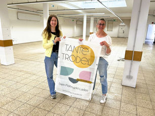 Zwei Frauen mit einem Banner in der Hand in einem leeren Ladengeschäft mit weißen Wänden.
