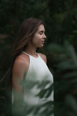 A young woman with long brown hair wearing a sleeveless white top stands in profile among green foliage, with soft natural light and a serene expression on her face meditating to enhance her psychic abilties.