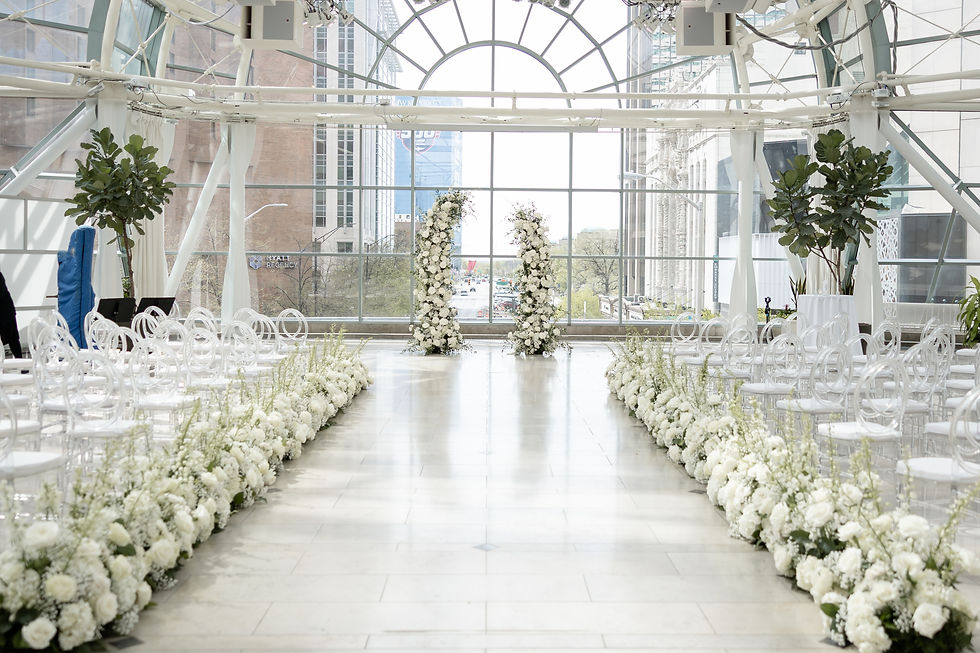 Wedding venue with white chairs and floral aisle in a glass-walled hall, city buildings visible outside, evoking elegance and anticipation.