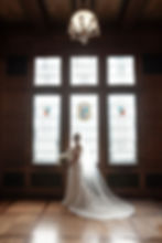 Bride in white gown with long veil holds a bouquet, standing by stained glass windows in elegant wooden room, beneath a chandelier.