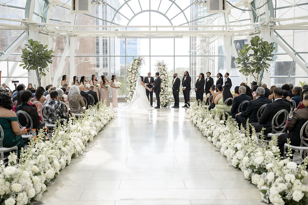 Bride and groom hold hands under floral arch in a sunlit venue. Bridesmaids in pink, groomsmen in suits, guests seated along flower-lined aisle.