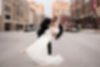 A couple dancing in a city street, the woman in a white dress. Joyful mood, surrounded by tall buildings and parked cars.