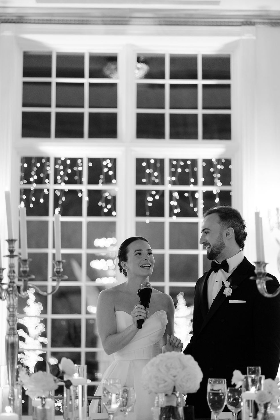 Bride and groom smiling, with the bride holding a microphone. They're in a decorated venue with lit candles and flowers, exuding joy.