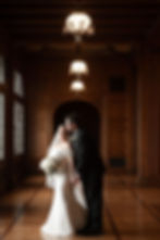 Bride in white gown holding bouquet, groom in black suit kisses her forehead. Elegant wood-paneled hall with chandeliers. Romantic mood.