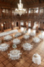 Elegant banquet hall with round tables, white linens, floral centerpieces, and gold chairs. A tiered cake on a separate table, under a grand chandelier.