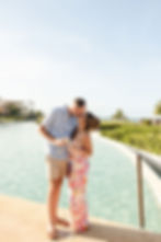 Couple kisses by a tropical resort pool, holding drinks, with palm trees and ocean in the sunny background.