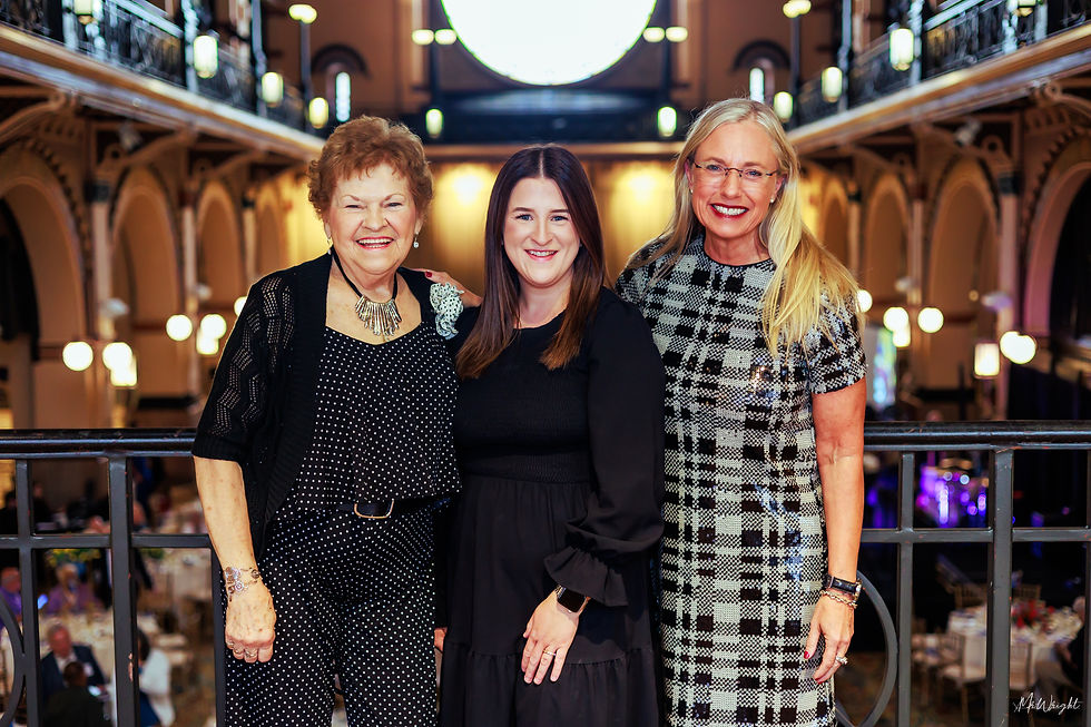 Three women smiling at the camera in an elegant, dimly lit venue with arched balconies. They wear black and checkered attire.