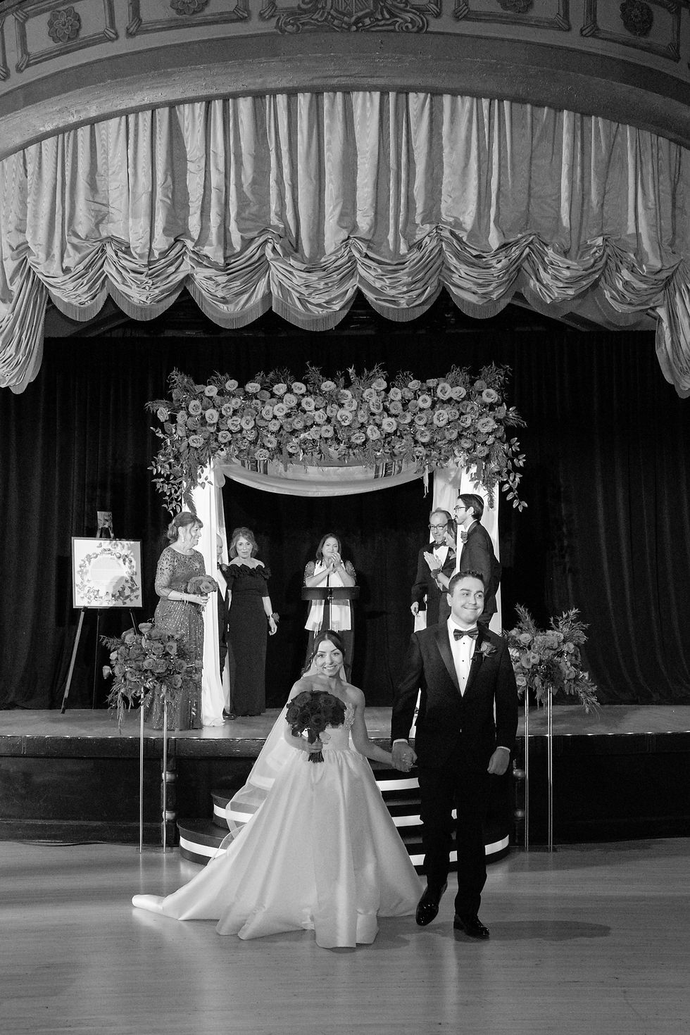 Bride in white gown and groom in black tuxedo walk down the aisle at a wedding ceremony under a floral arch, with guests in background.