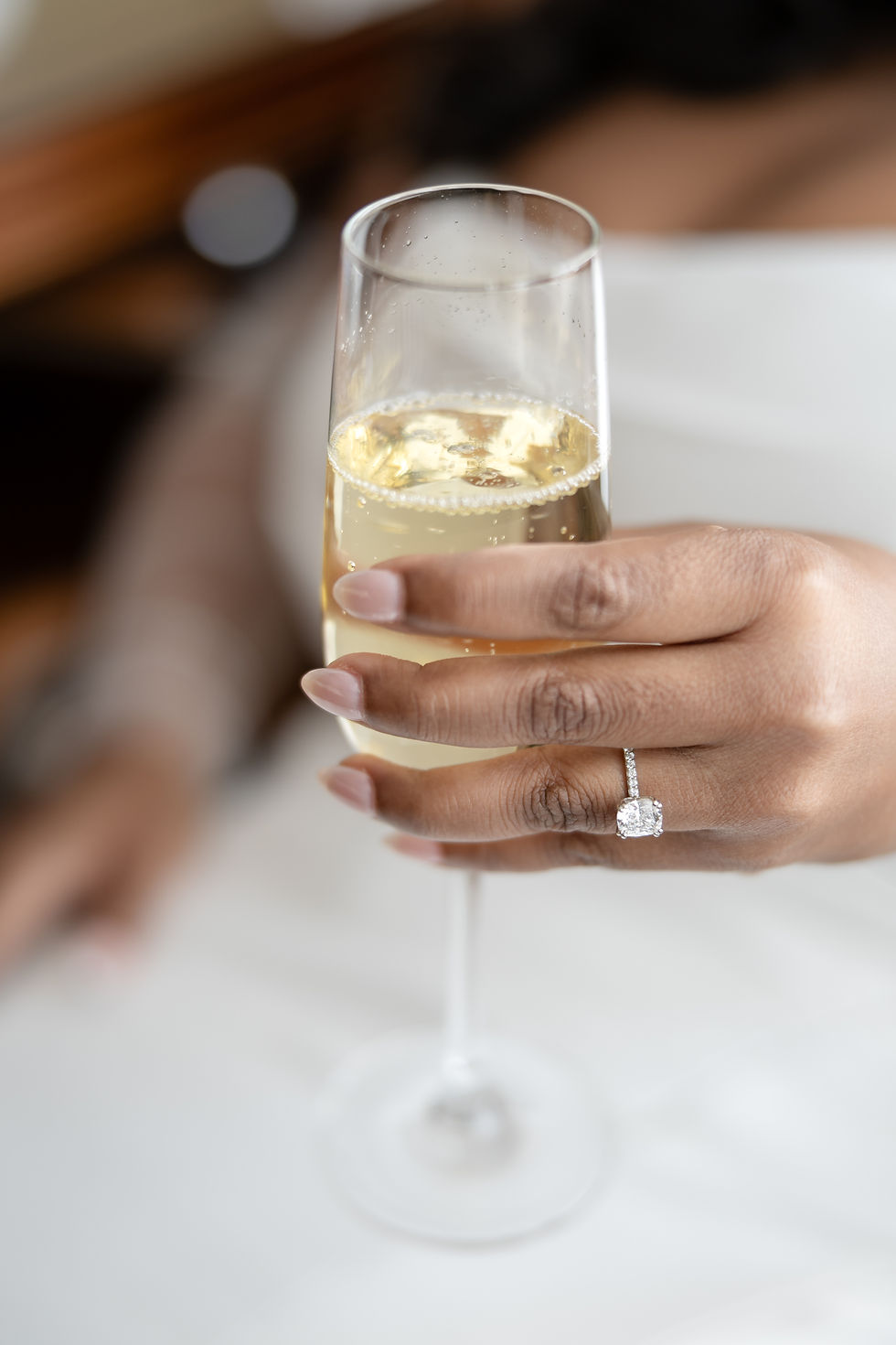 Hand holding a champagne flute, with a diamond ring visible. Background is softly blurred, creating an elegant and celebratory mood.