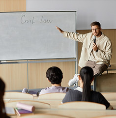 Adult male professor of law lecturing students while sitting on desk pointing at writing o
