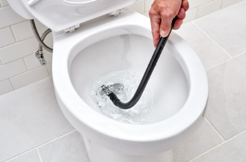 Hand using a black plumbing snake to unclog a white toilet in a tiled bathroom. The setting is clean and brightly lit.