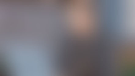 Cartoon man with glasses, wearing a gray shirt and apron, looks worried while typing on a laptop. Books and plants in background.