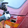 Person in gloves uses a power tool to repair a dent on a red car. Close-up of damage near the rear light in a sunny outdoor setting.