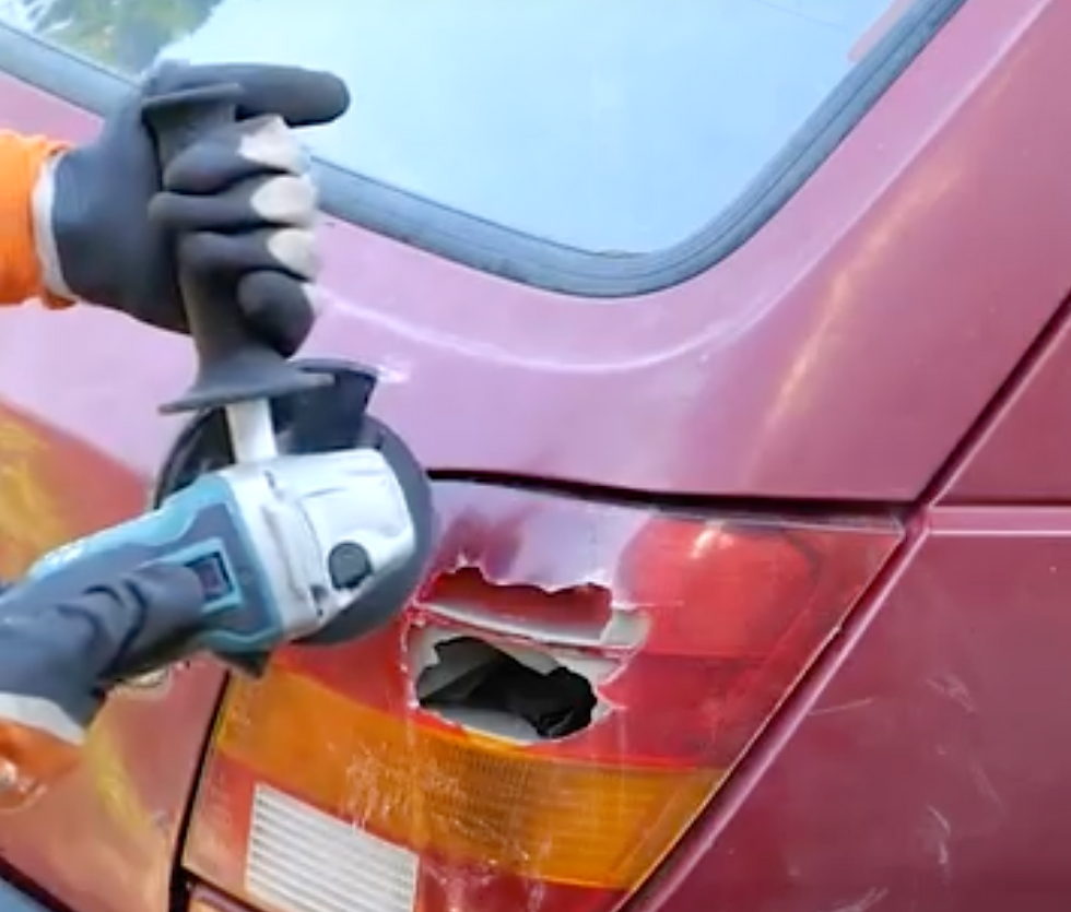 Person in gloves uses a power tool to repair a dent on a red car. Close-up of damage near the rear light in a sunny outdoor setting.