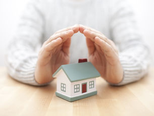 Hands forming a protective roof over a small house model on a table, symbolising home protection and security.