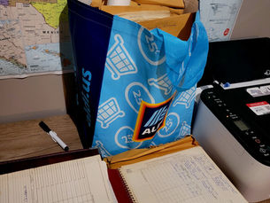 A large Aldi bag is pictured on a desk full of packets of genealogy work. A notebook is opened up in front of the bag displaying censuses.