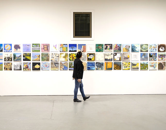 One person walks through a gallery looking at three rows of 12 x 12 flat art pieces hung in a white room. The person is facing to the right with their head turned away from the camera.