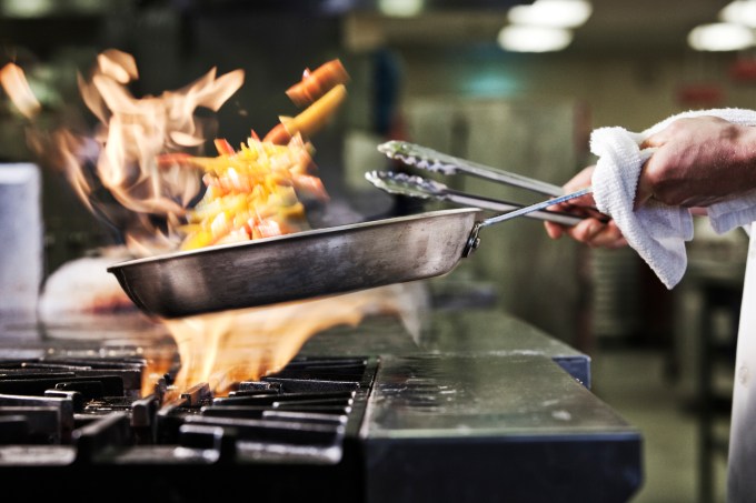 Close-up of chef's hands holding a sauté pan to cook food, flaming contents. Flames rising from the pan.
