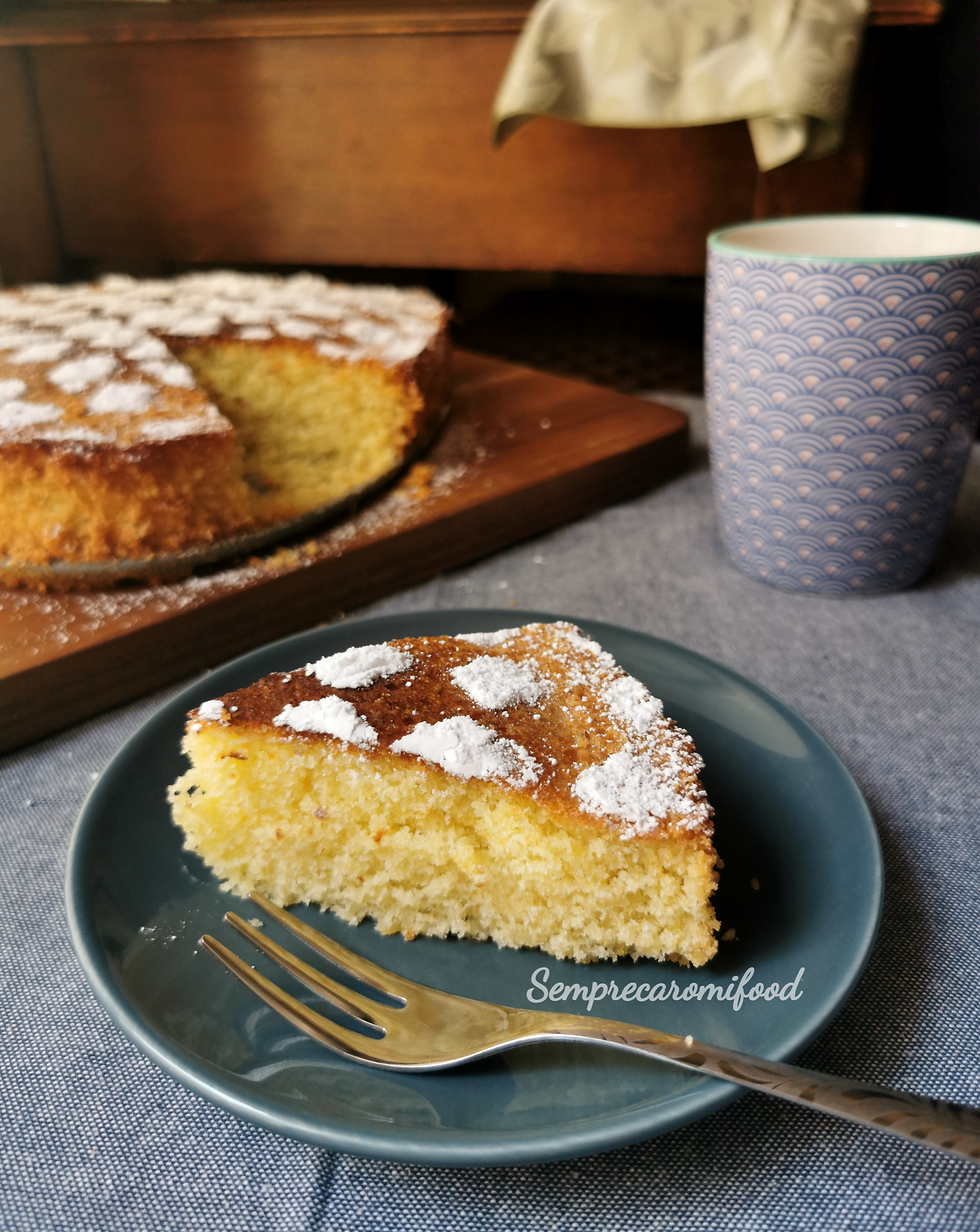 Torta fatta in casa con zucchero a velo, in primo piano una fetta su di un piattino grigio blu con tazza azzurra a motivi geometrici, su tovaglia azzurra e fondo legno. 
