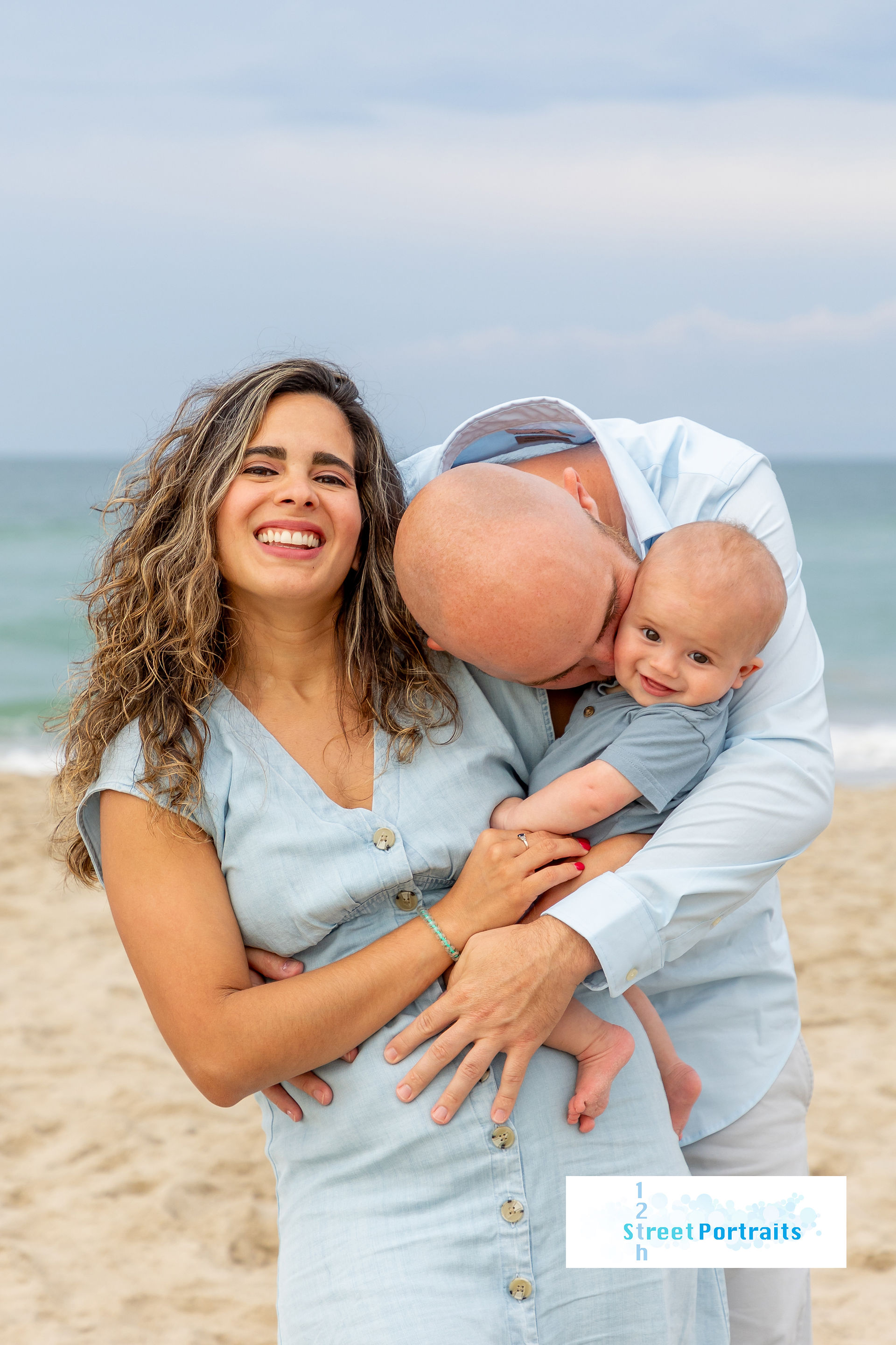 A mom and dad hold their baby. Dad is giving the baby a kiss
