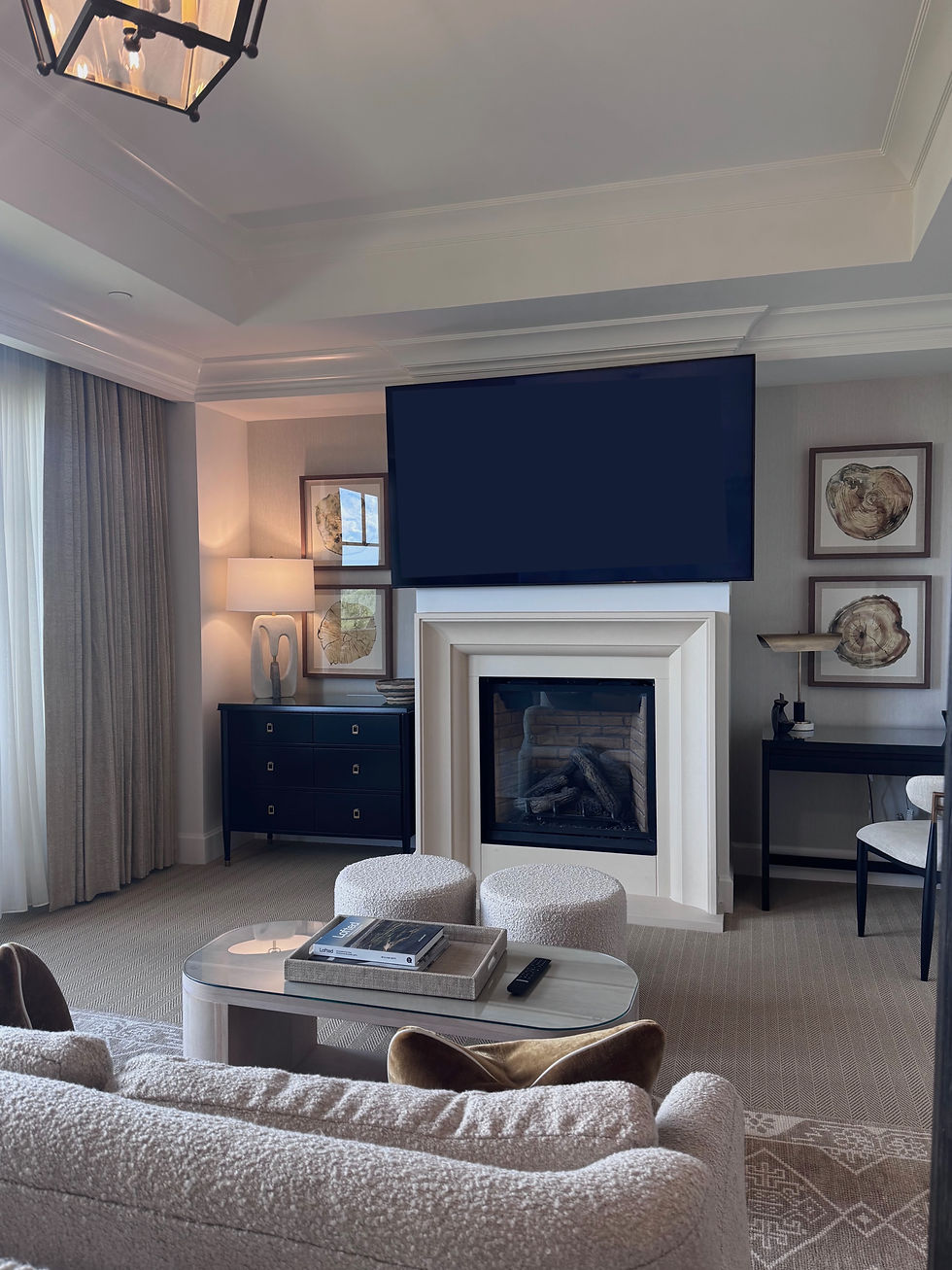 Elegant living room with a large TV above a fireplace, beige sofas, round stools, a black dresser, and framed art. Neutral tones create a calm mood.