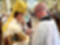 Bishop Heyd in a white chasuble, Gold Mitre. His is hold in crosier and hand is out stretched over an object praying. The object is held by a man with a shaved head and glasses, wearing a black cassock and white surplice.