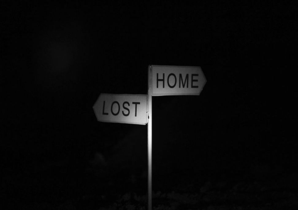 Wegweiser im Dunkeln mit zwei Schildern: "LOST" nach links, "HOME" nach rechts. Stimmung melancholisch, kontrastreiche Schwarz-Weiß-Optik.