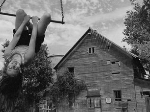A young girl hangs upside down as she swings from a tree swing in front of a barn.