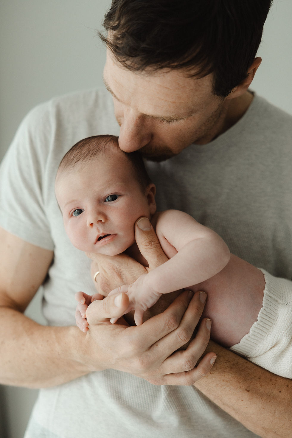 Father gently cradles a newborn baby, kissing its head. Both are in soft, neutral-toned clothing. The setting is calm and intimate.