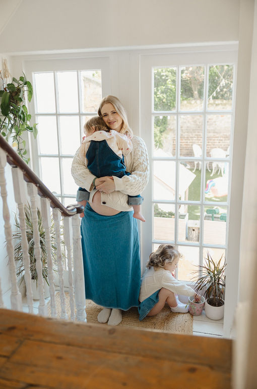 A pregnant woman cradling her toddler and baby bump while standing by a large, naturally lit window during a maternity photoshoot in London.