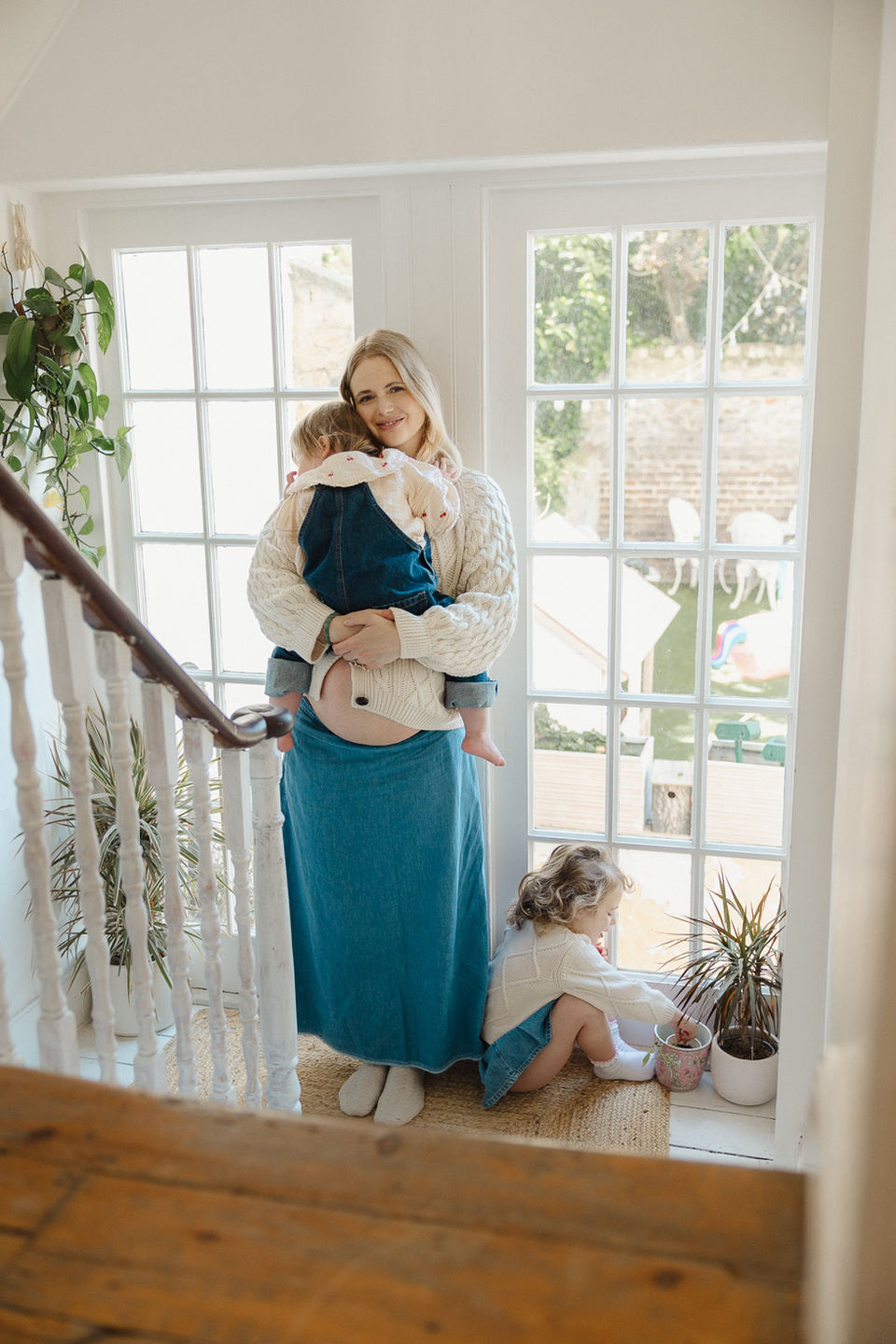 A pregnant woman cradling her toddler and baby bump while standing by a large, naturally lit window during a maternity photoshoot in London.