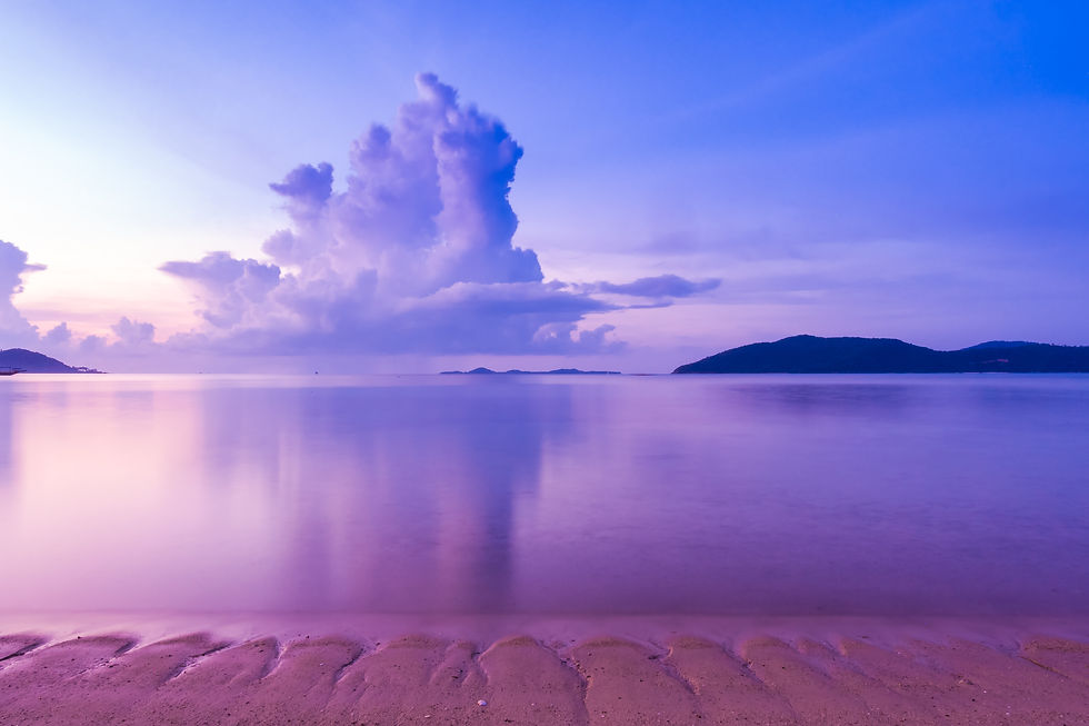 Calm beach at sunset with a vast purple sky and fluffy clouds. Serene water reflects colors. Distant hills on the horizon.