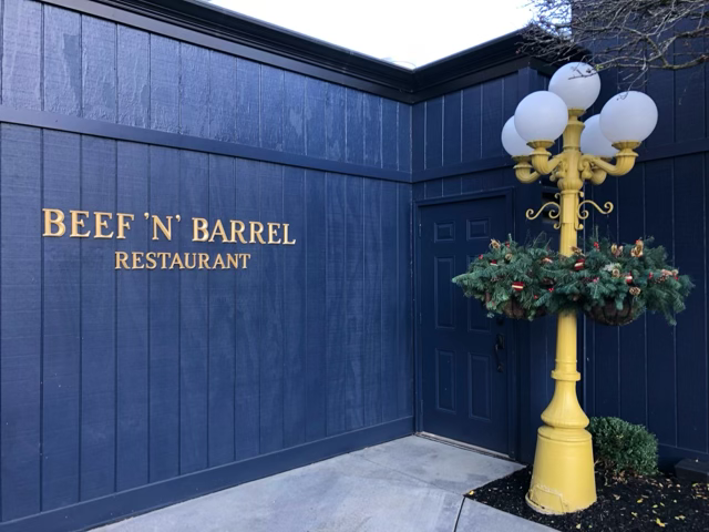 Rear entrance of Beef 'n' Barrel restaurant with blue exterior, gold signage and landscaping in Olean NY.