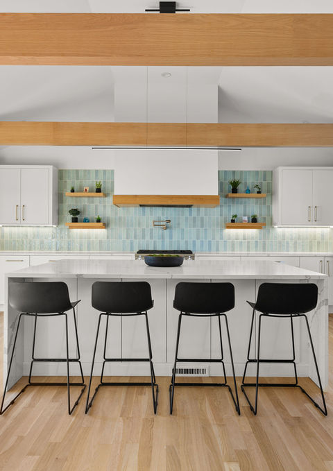 Full front view of a modern yet timeless kitchen in Fairfield, CT, showcasing a white quartz waterfall island with black chairs, white cabinetry with brass hardware, a gas range with sky-blue backsplash tiles, and light wood ceiling beams.
