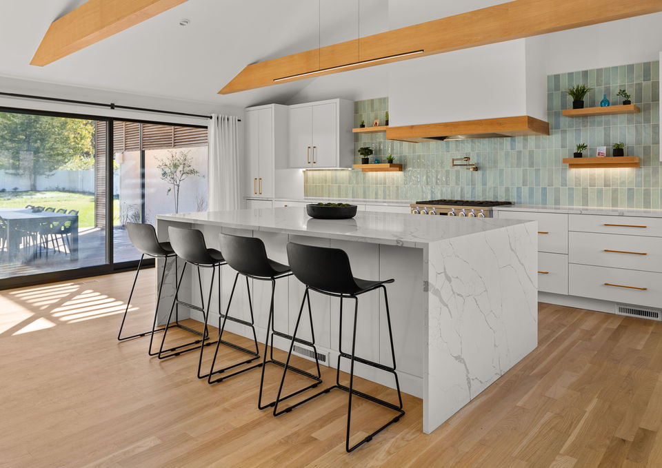 Side view of the modern kitchen in Fairfield, CT, featuring the white quartz island, gas range with blue backsplash, light wood beams, and large sliding glass doors opening to the custom outdoor patio.