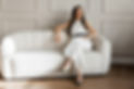 Anastasia Mitrovic, principal interior designer and founder serving Fairfield County, CT and Westchester, NY, sitting on a white couch in a white jumpsuit with a black belt, legs crossed, exuding elegance and professionalism.