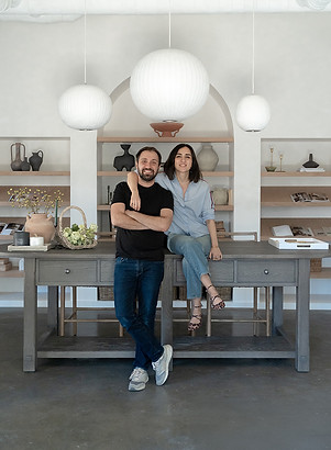 Alper and Burcu, founders of Shop by DNC, in their Atlanta-based design studio, with Alper leaning against a grey table and Burcu beside him, surrounded by styled built-ins featuring the curated products they sell.
