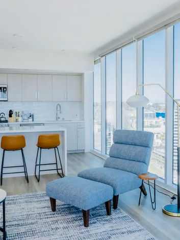 A sleek and modern living area featuring a blue couch, blue chaise lounge chair, and a white kitchen with floor-to-ceiling windows that allow for an abundance of natural light to pour in, creating a bright and airy atmosphere."