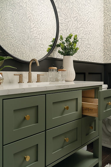 Bathroom design with green vanity, white quartz top, brass hardware, circular mirror, black wainscoting, and wallpaper, part of a Michigan luxury home renovation.