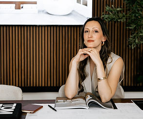 Anastasia Mitrovic, interior designer serving Connecticut and Manhattan, sitting at a design table, smiling at the camera with her hands under her chin, wearing a brown dress and a gold bracelet, exuding a professional yet approachable vibe in a stylish design workspace.