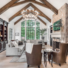 Open concept living area featuring wood beams on the ceiling, a grey couch, a cherry wood bookcase, a stone fireplace, and two leather chairs with throw blankets, creating a warm and inviting atmosphere.