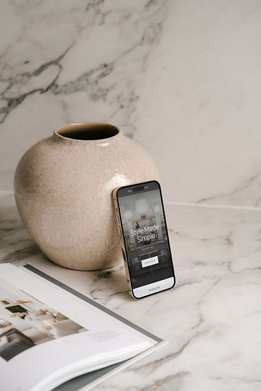 Mobile phone displaying the homepage of Shop by DNC’s custom Shopify website, leaning against a brown round vase on a marble kitchen counter, styled to reflect the clean and curated aesthetic of the brand.