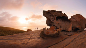 Rugged rock formations of Remarkable Rocks overlook a sunset-lit ocean. Two people walk along the cliff, sky adorned with clouds. A serene and awe-inspiring scene.