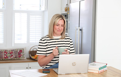 Anna Noden, compassionate therapist, smiling warmly at an open laptop while holding a cup of tea in a bright and welcoming kitchen.