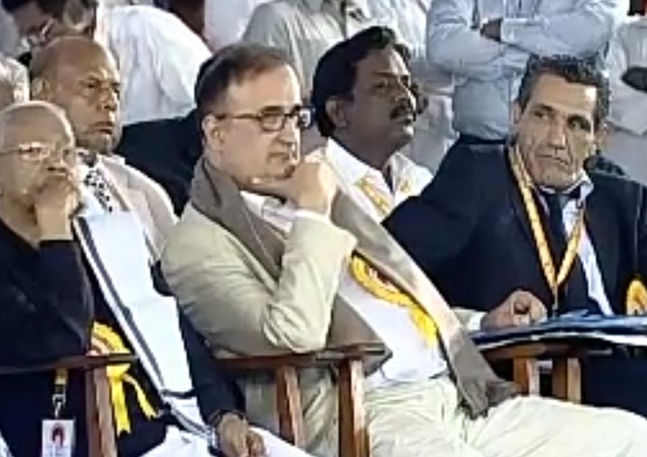 Man in glasses observing at formal event, part of Visual Diplomatic Registry.