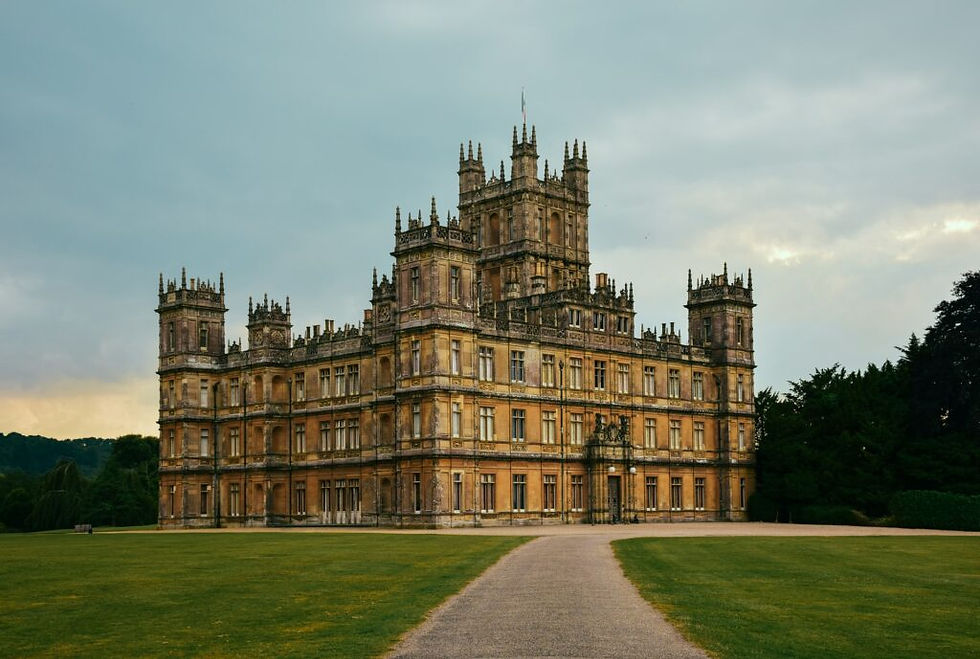 Grand historic castle with intricate towers set in lush green grounds under a cloudy sky. A winding path leads to the entrance.