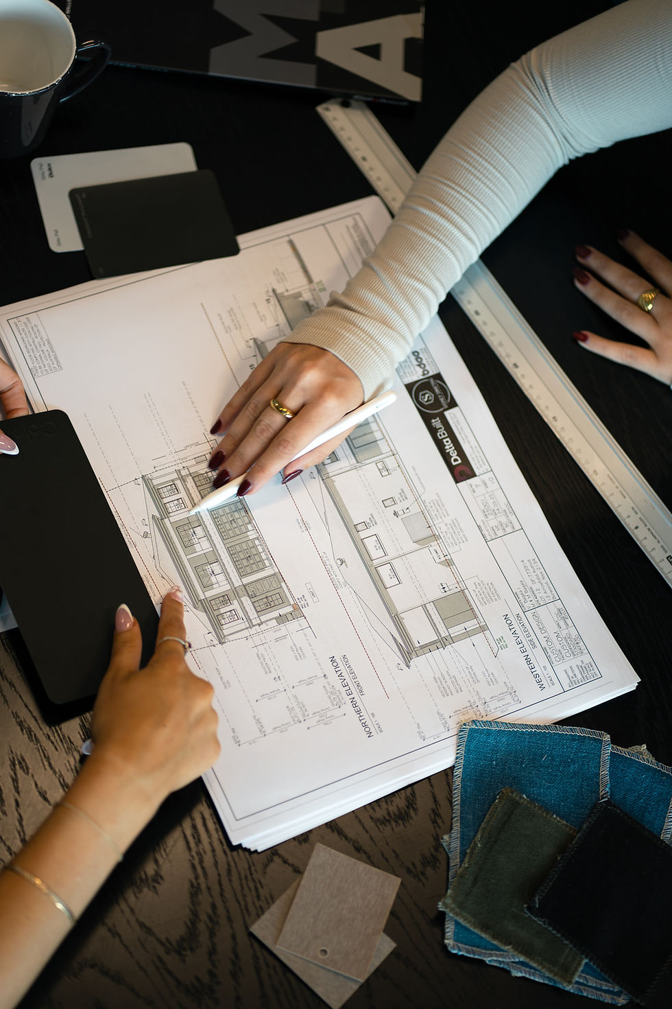Two people reviewing architectural blueprints on a table, with one person pointing at a building design while another holds a pen. Nearby are a tablet, material samples, rulers, and a coffee cup.